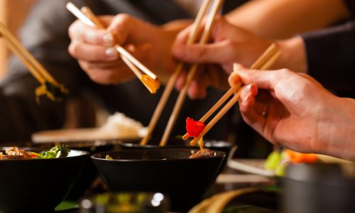 Close-up of hands using chopsticks to pick up food from small black bowls at a dining table.